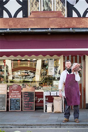 fresh beef displays - Man wearing apron standing on pavement outside butcher shop, carrying large piece of beef on his shoulder. Stock Photo - Premium Royalty-Free, Code: 6118-09148201