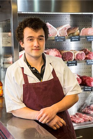 simsearch:6118-08729219,k - Man with brown curly hair wearing apron standing in butcher shop, smiling at camera. Foto de stock - Sin royalties Premium, Código: 6118-09148202