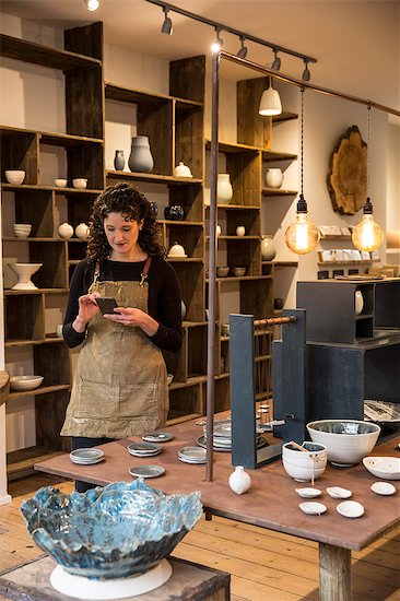 Woman with curly brown hair wearing apron standing in her pottery shop, looking at mobile phone. Stock Photo - Premium Royalty-Free, Image code: 6118-09148232