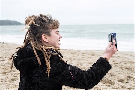 simsearch:6118-08023790,k - Smiling young woman with brown hair and dreadlocks and a lip piercing wearing black furry jacket, sitting on sandy beach by ocean, taking selfie with mobile phone. Stock Photo - Premium Royalty-Free, Code: 6118-09165843