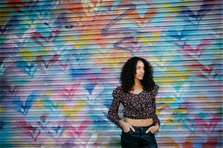 simsearch:6118-09039238,k - Young woman with black curly hair standing in front of shutter covered in colourful graffiti. Stock Photo - Premium Royalty-Free, Code: 6118-09165938