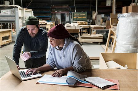 A Caucasian male carpenter and a Black female carpenter working on a laptop computer after work hours in a large woodworking factory. Stock Photo - Premium Royalty-Free, Code: 6118-09166024