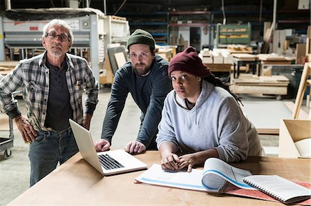 A portrait of mixed race carpenters around a laptop computer after work hours in a large woodworking facility. Stock Photo - Premium Royalty-Free, Code: 6118-09166025