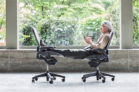 senior man feet up - An Asian businessman checking his phone, sitting in a chair and resting his feet in another chair. Stock Photo - Premium Royalty-Free, Code: 6118-09166195