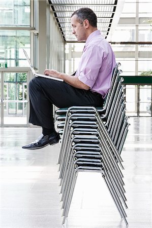 simsearch:6118-09173924,k - A Caucasian businessman sitting on top of multiple stacked chairs and working on a laptop computer. Stockbilder - Premium RF Lizenzfrei, Bildnummer: 6118-09166242