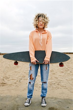 redondo beach - Young woman with curly blond hair wearing pink hoodie and ripped jeans standing on sandy beach, holding skateboard, looking at camera. Photographie de stock - Premium Libres de Droits, Code: 6118-09166307