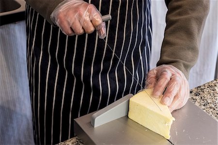 High angle close up of man wearing apron standing at counter in a delicatessen, slicing cheese with wire cutter. Photographie de stock - Premium Libres de Droits, Code: 6118-09173448