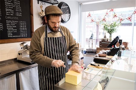 Bearded man wearing flat cap and apron standing at counter in a delicatessen, cutting cheese. Photographie de stock - Premium Libres de Droits, Code: 6118-09173449
