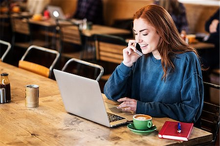 simsearch:649-08924663,k - Smiling young woman with long red hair sitting at table, working on laptop computer, using mobile phone. Foto de stock - Royalty Free Premium, Número: 6118-09173403