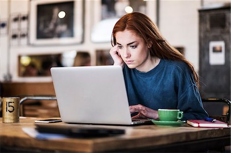 simsearch:649-08924663,k - Young woman with long red hair sitting at table, working on laptop computer. Foto de stock - Royalty Free Premium, Número: 6118-09173401