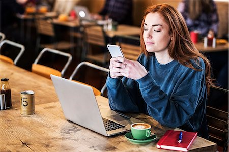 Smiling young woman with long red hair sitting at table, working on laptop computer, using mobile phone. Foto de stock - Royalty Free Premium, Número: 6118-09173402