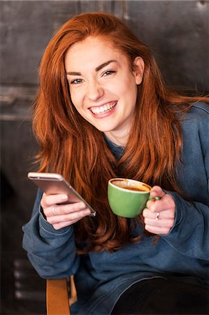 Smiling young woman with long red hair sitting at table, holding mobile phone and cup of coffee, looking at camera. Foto de stock - Royalty Free Premium, Número: 6118-09173412