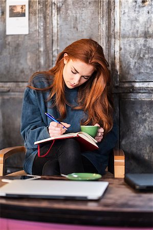 Young woman with long red hair sitting at table, holding notebook and cup of coffee. Foto de stock - Royalty Free Premium, Número: 6118-09173414
