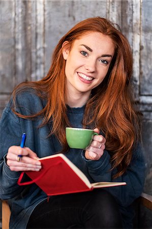 Smiling young woman with long red hair sitting at table, holding notebook and cup of coffee, looking at camera. Foto de stock - Royalty Free Premium, Número: 6118-09173416
