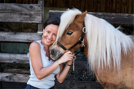 Smiling woman standing outside stable, holding a Shetland pony with a long blonde mane by its bridle. Photographie de stock - Premium Libres de Droits, Code: 6118-09173731