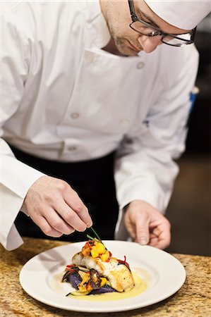 A Caucasian male chef putting the finishing touches on a plate of fish in a commercial kitchen. Photographie de stock - Premium Libres de Droits, Code: 6118-09173956