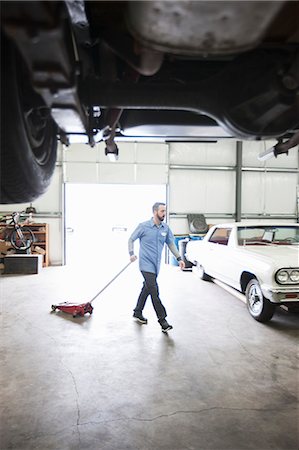 restoring car in garage - A male car mechanic pulls a portable car jack across the floor of the car repair shop. Stock Photo - Premium Royalty-Free, Code: 6118-09173994