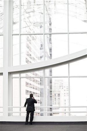 simsearch:6118-09173926,k - A businessman talking on a cell phone while standing in front of a window in a large convention centre lobby. Stock Photo - Premium Royalty-Free, Code: 6118-09173925