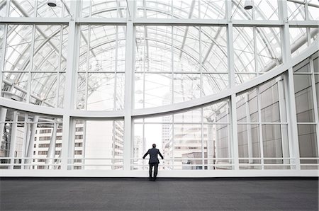simsearch:6118-09173926,k - A man standing in an open space in a glass atrium in an office building, leaning on a railing, rear view. Stock Photo - Premium Royalty-Free, Code: 6118-09173933