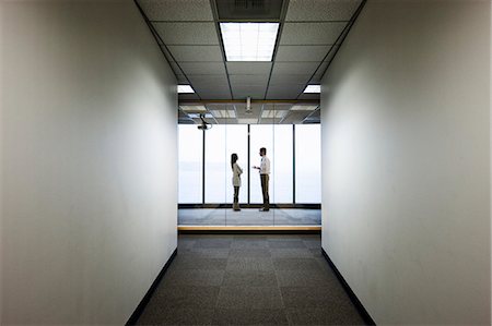 Male and female business people talking and standing in front of a window at the end of a long hallway. Stock Photo - Premium Royalty-Free, Code: 6118-09174295