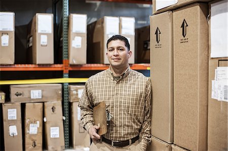 simsearch:6118-09174228,k - A Hispanic male warehouse worker standing next to racks of boxed products in a distribution warehouse. Fotografie stock - Premium Royalty-Free, Codice: 6118-09174210