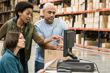 A group of three mixed race warehouse workers checking inventory on a computer in a large distribution warehouse. Stock Photo - Premium Royalty-Free, Code: 6118-09174225