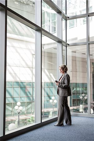 simsearch:6118-09174347,k - A Caucasian businesswoman checking her cell phone for messages while standing next to a large window in a convention centre lobby. Fotografie stock - Premium Royalty-Free, Codice: 6118-09174370