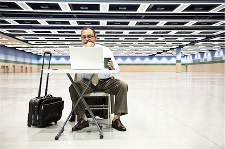 simsearch:6118-09174347,k - A Hispanic businessman looking at a laptop computer sitting at a small desk in the middle of a convention arena. Fotografie stock - Premium Royalty-Free, Codice: 6118-09174377
