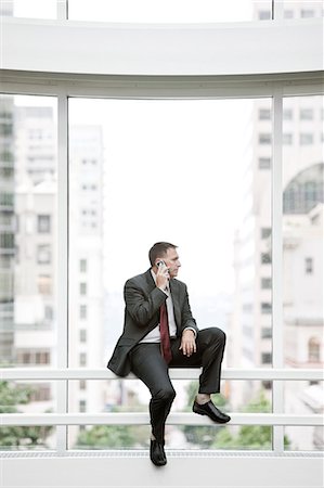 simsearch:6118-09174347,k - A Caucasian businessman using his phone while sitting by a railing at the base of a large window in a convention centre. Fotografie stock - Premium Royalty-Free, Codice: 6118-09174390