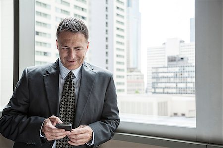 simsearch:6118-09174347,k - A Caucasian businessman texting on his phone while standing next to a window. Fotografie stock - Premium Royalty-Free, Codice: 6118-09174395
