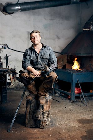simsearch:6118-09174461,k - Portrait of an artisan metalworker in his workshop, sitting on a wood block holding a long hammer. Stockbilder - Premium RF Lizenzfrei, Bildnummer: 6118-09174471