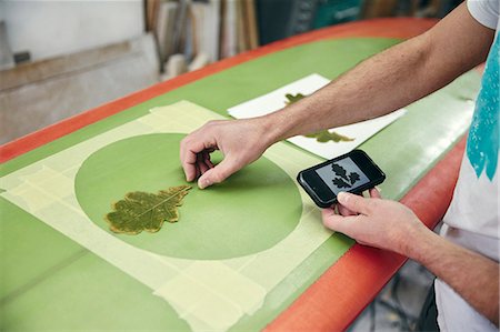 Man creating a design on surfboard laying an oak leaf on the board, using mobile phone in workshop Foto de stock - Sin royalties Premium, Código: 6118-09174474