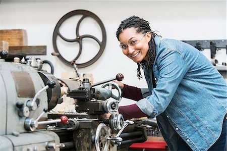 Smiling woman wearing safety glasses standing in a metal workshop at a machine, looking at camera. Stock Photo - Premium Royalty-Free, Code: 6118-09112055