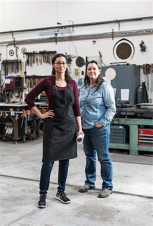 female artisan - Two women wearing apron and Denim shirt standing in metal workshop, smiling at camera. Stock Photo - Premium Royalty-Free, Code: 6118-09112003