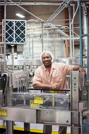 simsearch:6118-09129432,k - A portrait of an African American worker wearing a head net next to a conveyor belt of lemon flavoured water in a bottling plant. Fotografie stock - Premium Royalty-Free, Codice: 6118-09129419