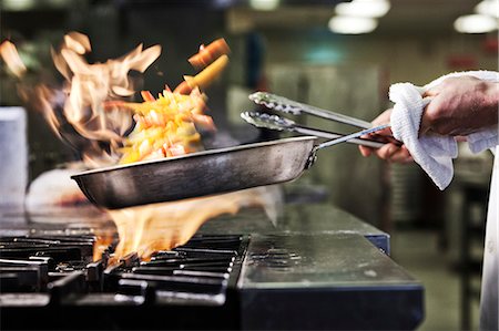 Close-up of chef's hands holding a sauté pan to cook food, flambéing contents. Flames rising from the pan. Stock Photo - Premium Royalty-Free, Code: 6118-09129746