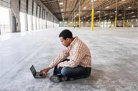 Black man working on lap top computer in front of loading dock doors in a new warehouse. Stock Photo - Premium Royalty-Free, Code: 6118-09139956