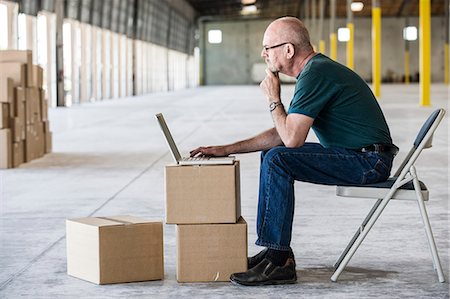 Caucasian man using boxes as desk and working on lap top computer in front of loading dock doors in a new warehouse. Stock Photo - Premium Royalty-Free, Code: 6118-09139958