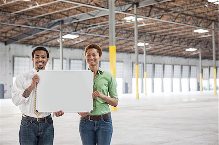 Black man and black woman holding up a blank slate in the interior of a brand new empty warehouse space. Stock Photo - Premium Royalty-Free, Code: 6118-09139933