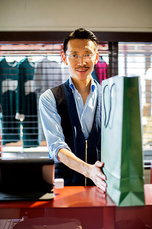 Japanese salesman with moustache wearing glasses standing at counter in clothing store, holding green shopping bag, smiling at camera. Stock Photo - Premium Royalty-Free, Code: 6118-09200222