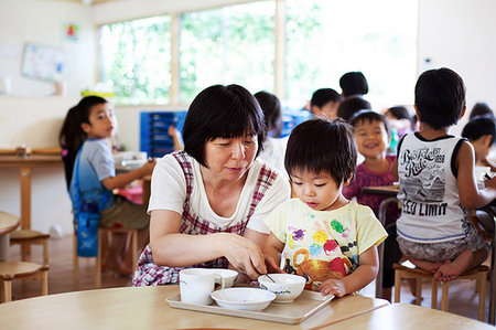 Female teacher sitting at table in a Japanese preschool, helping young boy to eat his lunch. Stock Photo - Premium Royalty-Free, Code: 6118-09200518