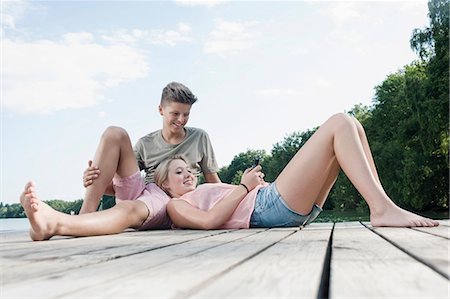 pantalones cortos - Teenage couple with smartphone relaxing on a jetty at lake Foto de stock - Sin royalties Premium, Código: 6121-07741786