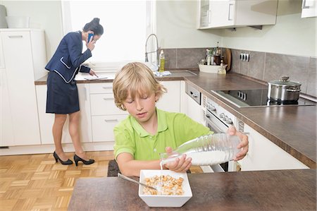 Boy pouring milk in muesli while mother has business call in kitchen, Bavaria, Germany Foto de stock - Sin royalties Premium, Código: 6121-08859177