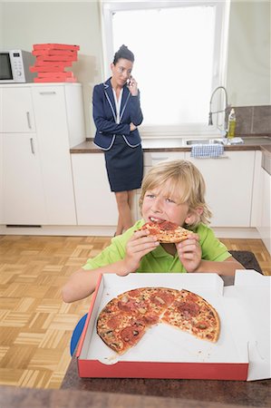 front - Boy eating pizza while mother has business call in kitchen, Bavaria, Germany Stock Photo - Premium Royalty-Free, Code: 6121-08859178