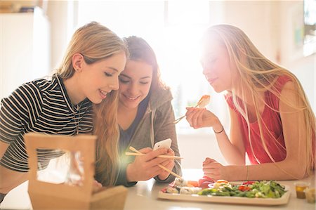 Teenage girls eating sushi and texting with cell phone in sunny kitchen Stock Photo - Premium Royalty-Free, Code: 6124-08170481