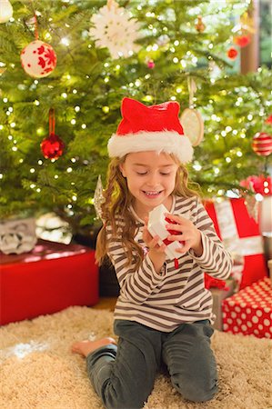 simsearch:6108-05874752,k - Smiling girl in Santa hat opening gift in front of Christmas tree Photographie de stock - Premium Libres de Droits, Code: 6124-08926896