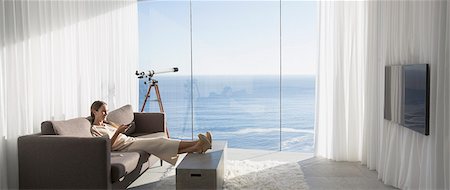 simsearch:632-02744819,k - Woman relaxing with feet up, watching TV in modern, luxury home showcase interior living room with sunny ocean view Foto de stock - Sin royalties Premium, Código: 6124-09099765