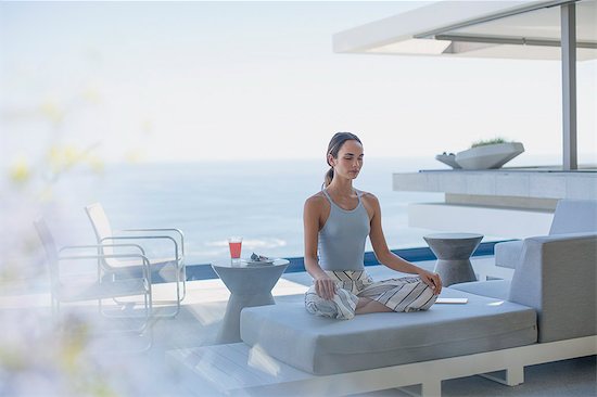 Serene woman meditating in lotus position on modern, luxury home showcase exterior patio sofa with ocean view Stock Photo - Premium Royalty-Free, Image code: 6124-09099809