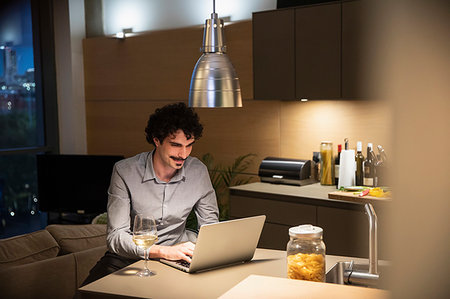 Man drinking white wine at laptop in apartment kitchen at night Photographie de stock - Premium Libres de Droits, Code: 6124-09239399