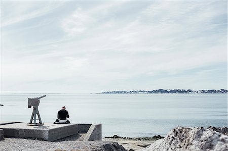 Rear view of man sitting by telescope overlooking sea against sky Photographie de stock - Premium Libres de Droits, Code: 6127-08666694